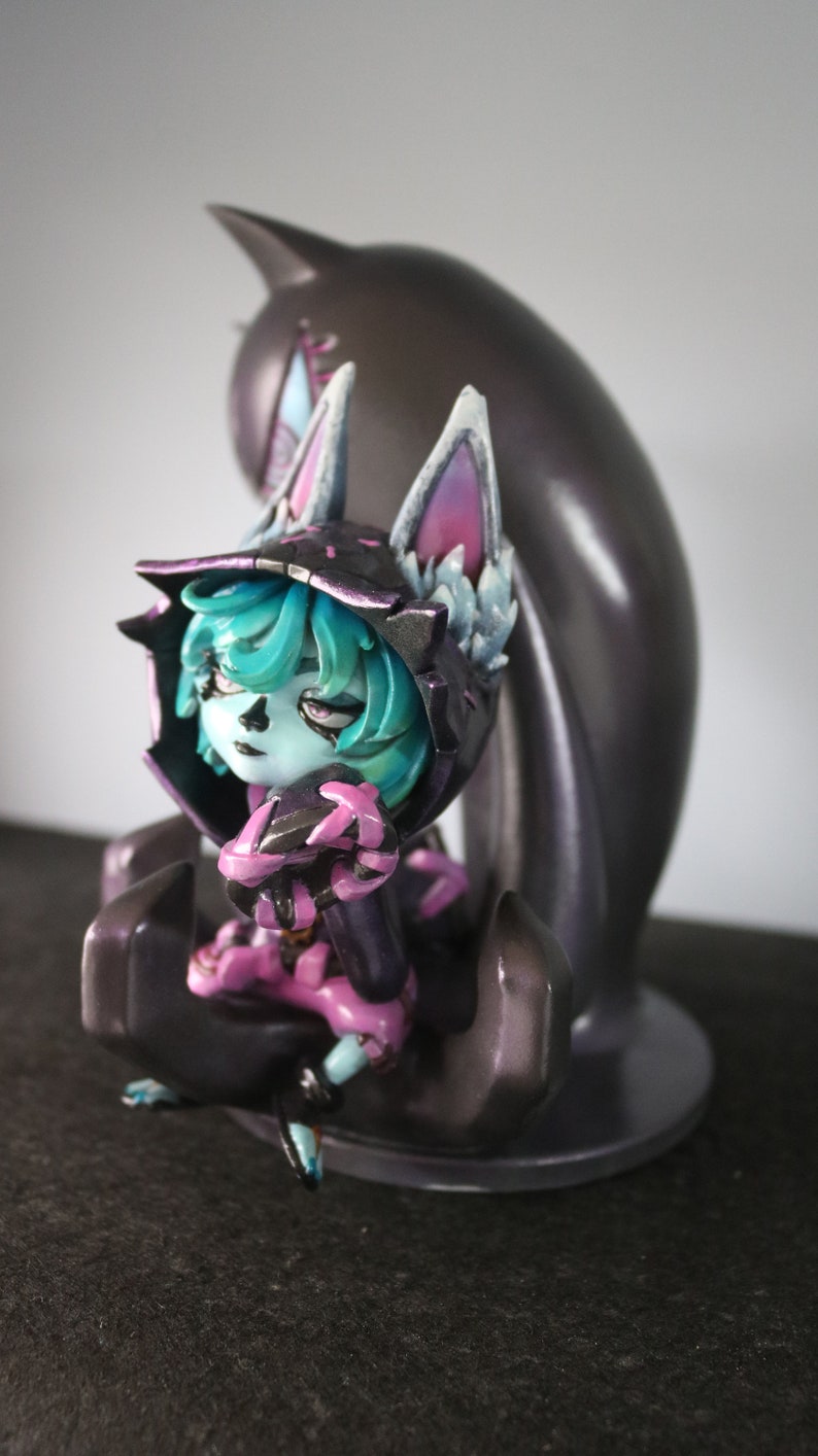 May include: A detailed figurine of a character with teal hair, a black hooded cloak, and pink accents. The figure is seated in a stylized pose, with a dark, amorphous shape behind it. The figurine is on a dark surface.