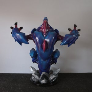 May include: A blue and red fantasy creature figurine with outstretched arms. The figure has a detailed, textured design and is mounted on a base of gray, crystalline structures. The color scheme includes shades of blue, red, and black.