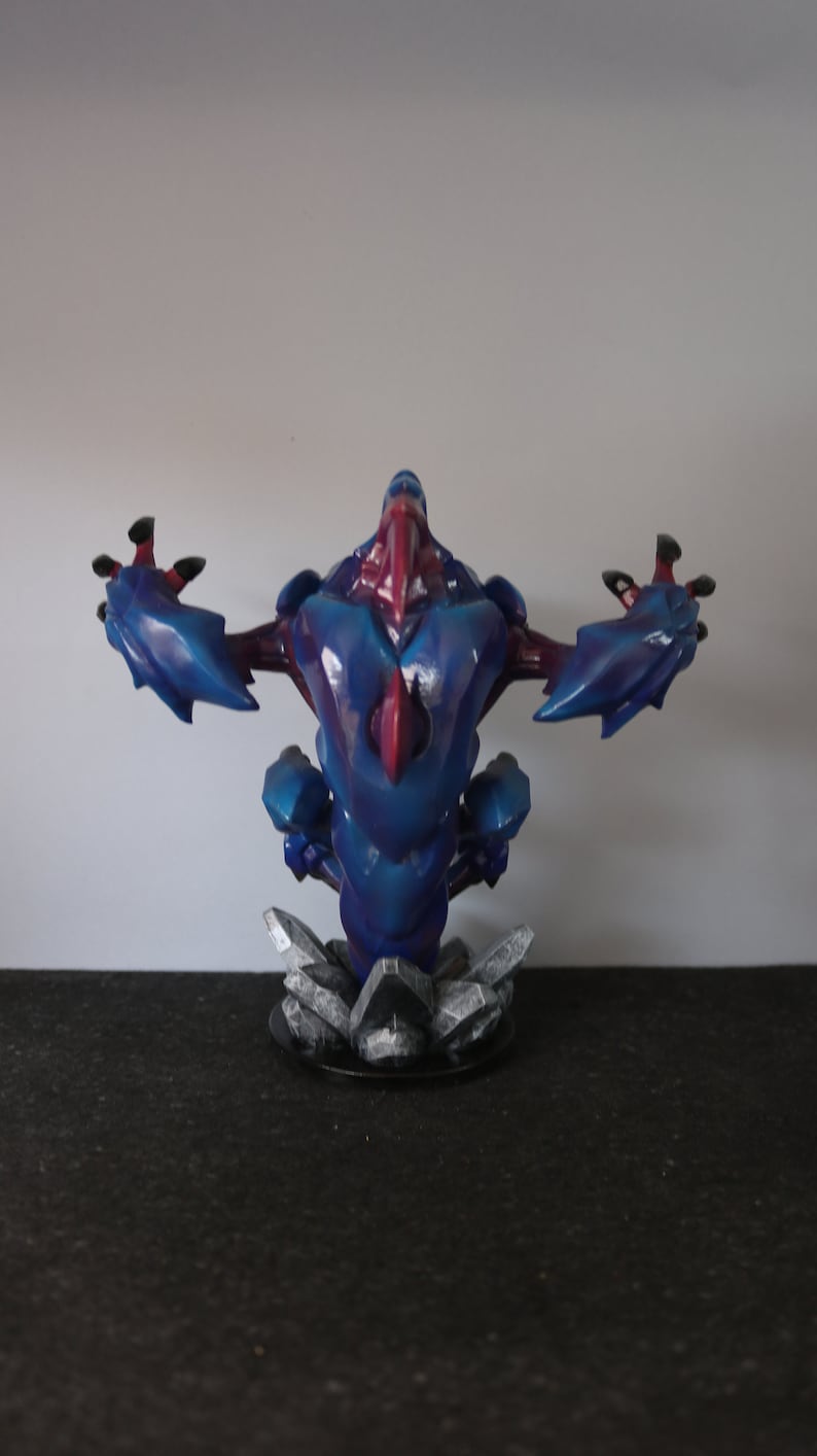 May include: A blue and red fantasy creature figurine with outstretched arms. The figure has a detailed, textured design and is mounted on a base of gray, crystalline structures. The color scheme includes shades of blue, red, and black.