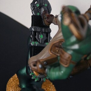 Pu&ograve; includere: Statuetta dettagliata di un personaggio di fantascienza con armatura verde e marrone, in posa vicino a una struttura scura e futuristica. La base &egrave; strutturata con un aspetto sabbioso e la struttura ha accenti verdi luminosi.