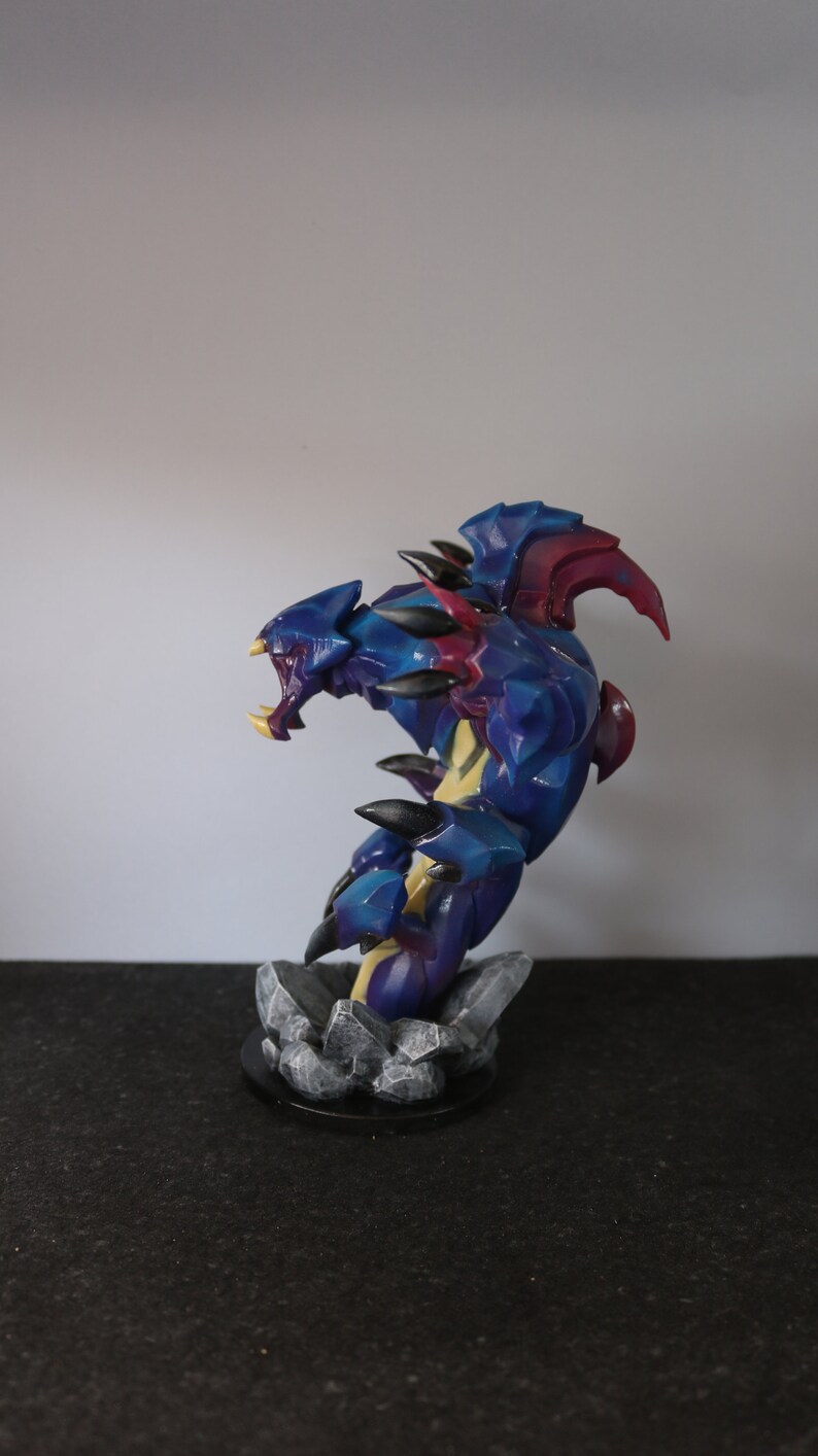 May include: A detailed figurine of a fantasy creature with a blue and purple color scheme. The figure has sharp claws, teeth, and spikes. It is posed on a rocky base, with a black circular base. The figurine is approximately 6 inches tall.
