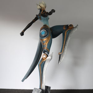 May include: A detailed figurine of a female character in a dynamic pose. The figure is predominantly teal and gold, with silver accents. It stands on a dark grey base with rock-like details, showcasing a blend of fantasy and artistry.