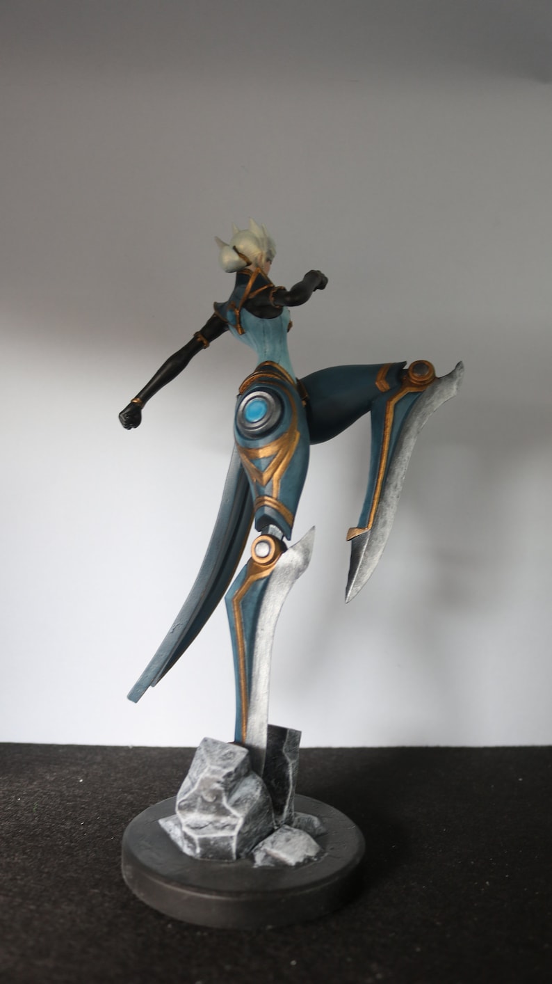 May include: A detailed figurine of a female character in a dynamic pose. The figure is predominantly teal and gold, with silver accents. It stands on a dark grey base with rock-like details, showcasing a blend of fantasy and artistry.