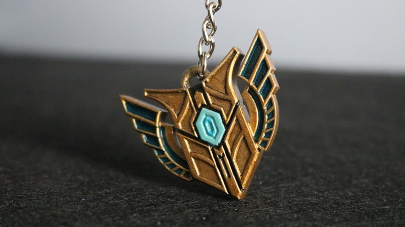 Op de afbeelding: Een goudkleurige metalen sleutelhangerhanger met teal accenten en een centrale teal-kleurige edelsteen. De hanger heeft een gestileerd ontwerp met vleugelachtige elementen en is bevestigd aan een zilveren ketting. Het algehele ontwerp is geometrisch en symmetrisch.