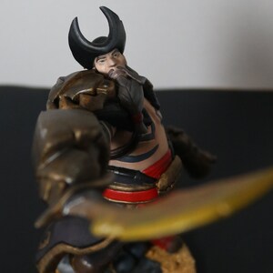 May include: A detailed figurine of a warrior, featuring a horned helmet and ornate armor. The figure holds a golden blade, with intricate details on the clothing and face. The color palette includes gold, black, red, and brown.