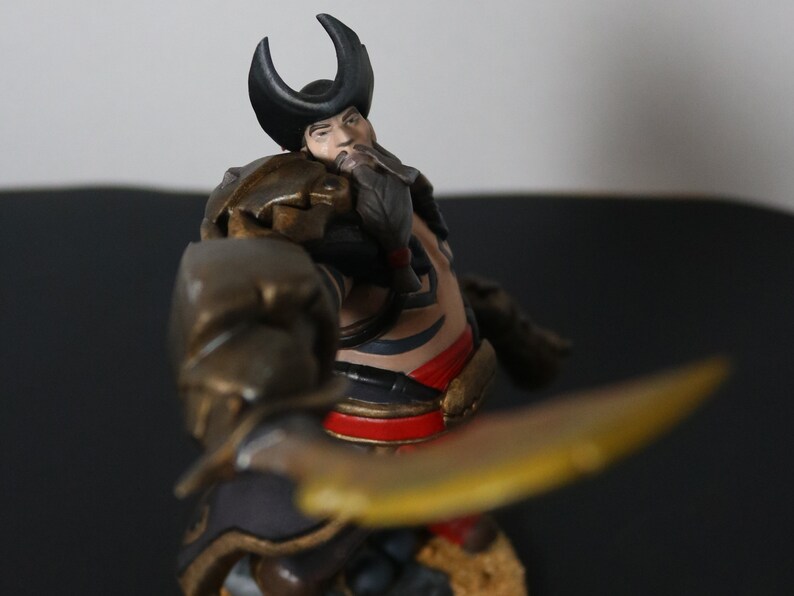 May include: A detailed figurine of a warrior, featuring a horned helmet and ornate armor. The figure holds a golden blade, with intricate details on the clothing and face. The color palette includes gold, black, red, and brown.
