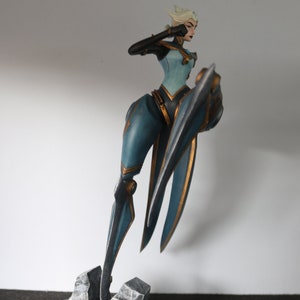 May include: A detailed figurine of a female character in a dynamic pose. The figure is clad in a teal and gold outfit, with a stylized shield. The base is a dark grey circle with textured rock formations. The character has white hair and is holding up her arm.