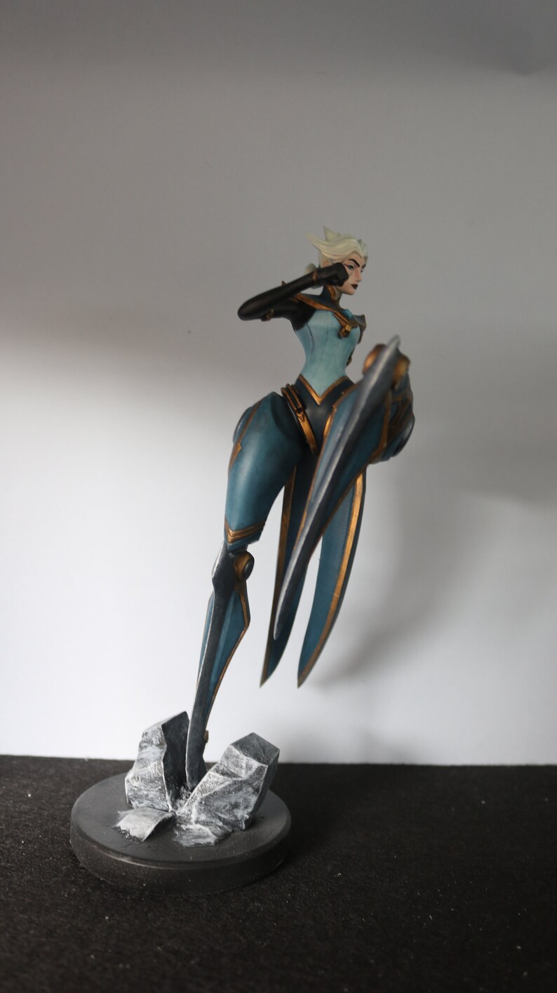 May include: A detailed figurine of a female character in a dynamic pose. The figure is clad in a teal and gold outfit, with a stylized shield. The base is a dark grey circle with textured rock formations. The character has white hair and is holding up her arm.