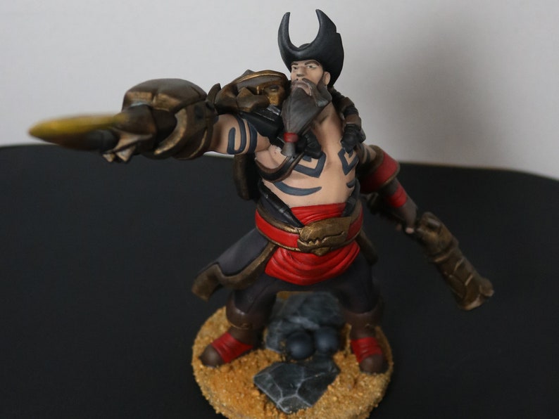 May include: A detailed figurine of a character with a beard, black hat, and tattoos. The figure is posed in a dynamic stance, wearing a red sash, black pants, and brown armor. The base is textured with sand and rocks.