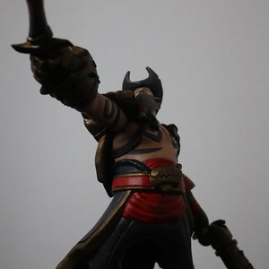 May include: A detailed figurine of a pirate, holding a curved blade and a pistol. The figure wears a tricorn hat, a red sash, and black pants. The base is textured with sand and small rocks. The color palette includes gold, red, black, and tan.