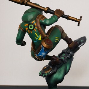 Pu&ograve; includere: Statuetta dettagliata di un personaggio in abito verde, in posa su un oggetto simile a uno snowboard. La figura tiene in mano un bastone. La base &egrave; nera con una texture dorata simile alla sabbia. La figura ha accenti marroni e un design dettagliato.