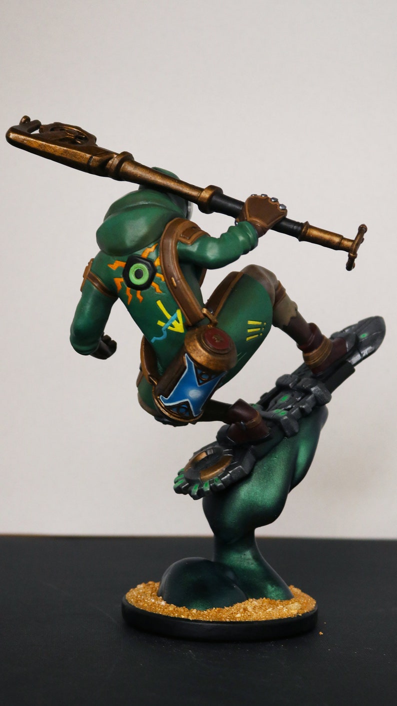 Pu&ograve; includere: Statuetta dettagliata di un personaggio in abito verde, in posa su un oggetto simile a uno snowboard. La figura tiene in mano un bastone. La base &egrave; nera con una texture dorata simile alla sabbia. La figura ha accenti marroni e un design dettagliato.