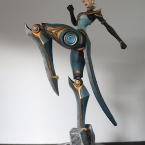 May include: A teal and gold figurine of a female character in a dynamic pose. The figure has black accents and blonde hair. The base is a dark grey, textured design. The character is in a fighting stance, with one leg raised.