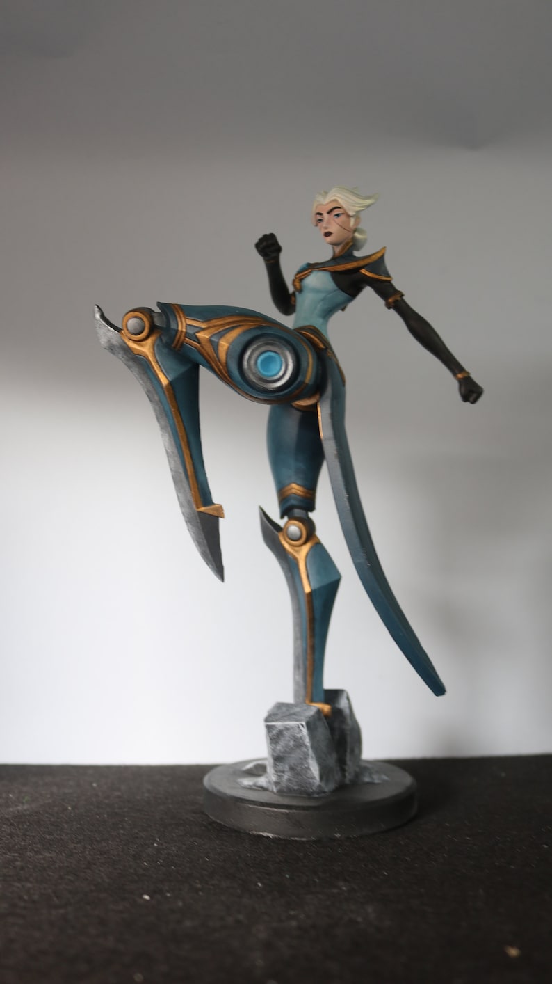 May include: A teal and gold figurine of a female character in a dynamic pose. The figure has black accents and blonde hair. The base is a dark grey, textured design. The character is in a fighting stance, with one leg raised.