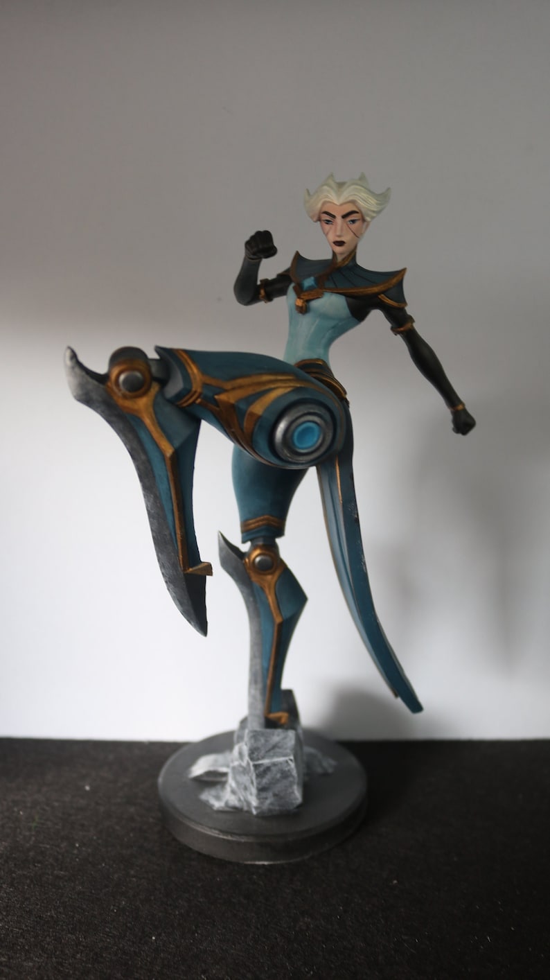May include: A detailed figurine of a female character in a dynamic pose. The figure is predominantly teal and gold, with a stylized design. The base is a dark grey circle with a rocky texture. The character has white hair and a determined expression.