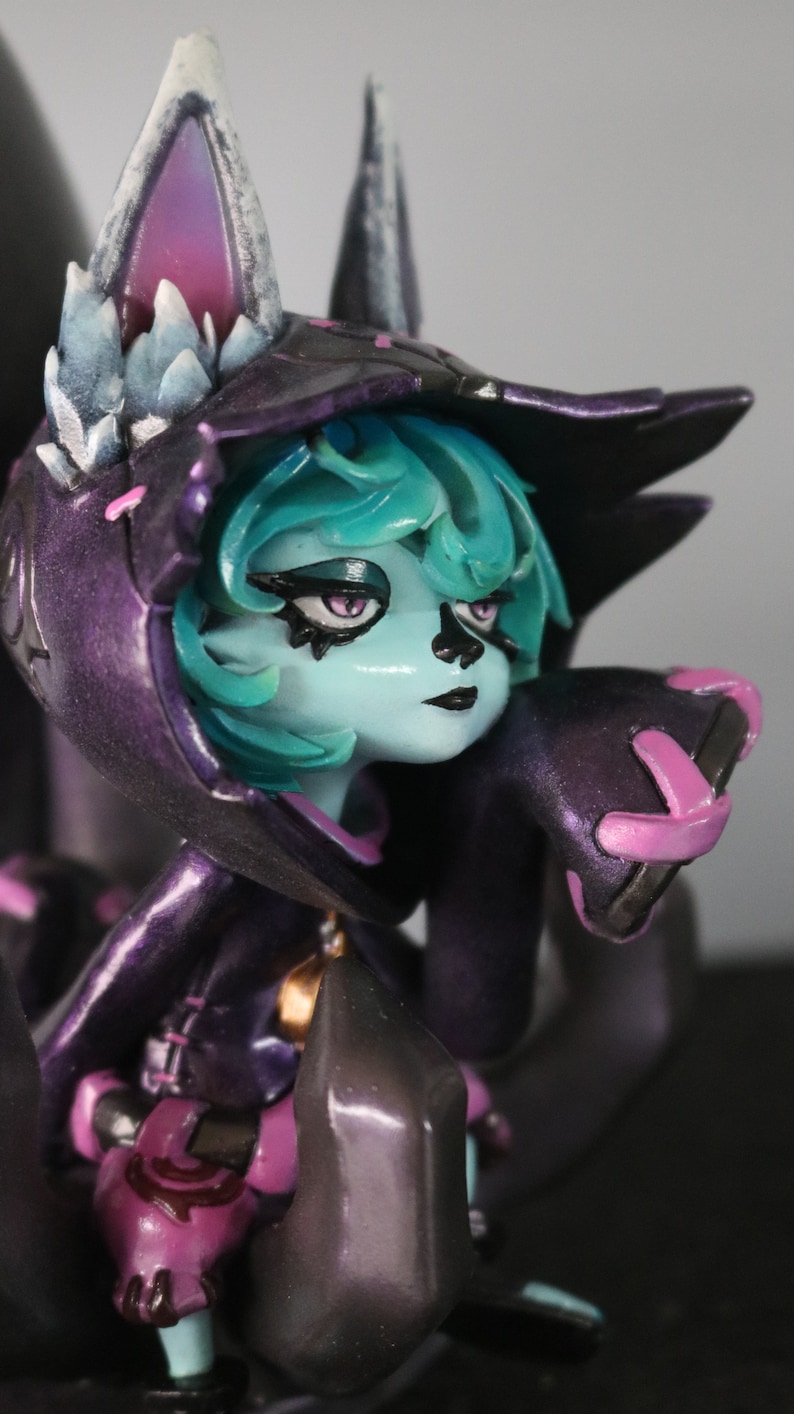 May include: A detailed figurine of a character with teal hair and a purple hooded cloak. The figure has a pale blue face with dark eye makeup and black lips. The cloak has pink accents and the figure is holding a dark grey object.