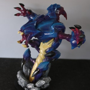 May include: A detailed figurine of a creature with a blue and purple exoskeleton, yellow underbelly, and sharp claws. The figure is posed on a rocky base, showcasing intricate details and a dynamic design. The creature has an open mouth with sharp teeth.