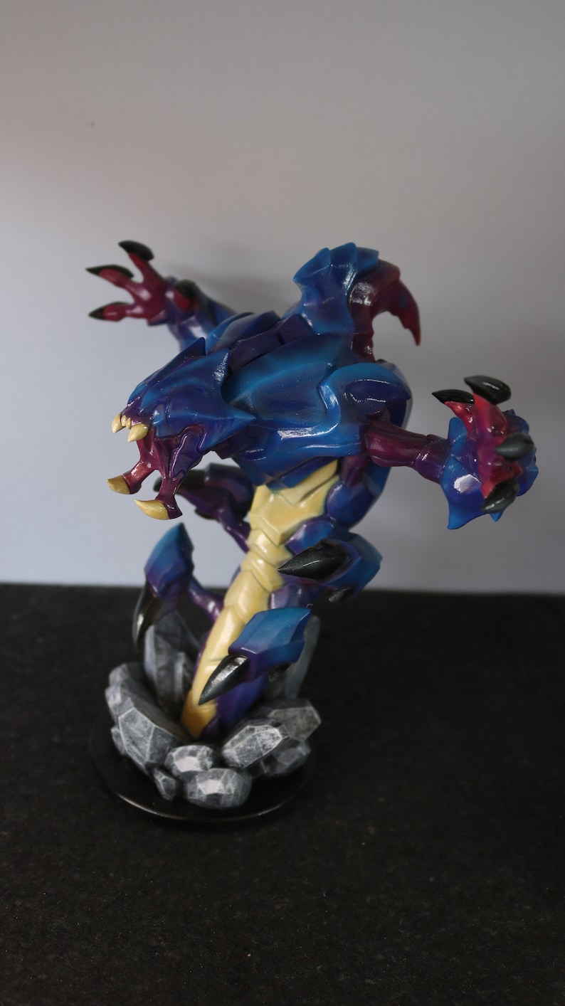 May include: A detailed figurine of a creature with a blue and purple exoskeleton, yellow underbelly, and sharp claws. The figure is posed on a rocky base, showcasing intricate details and a dynamic design. The creature has an open mouth with sharp teeth.