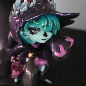 May include: A detailed figurine of a character with a fox-like hood and ears. The figure has teal hair, a pale blue face, and is dressed in a dark purple outfit with pink accents. The ears are white with purple and blue details.