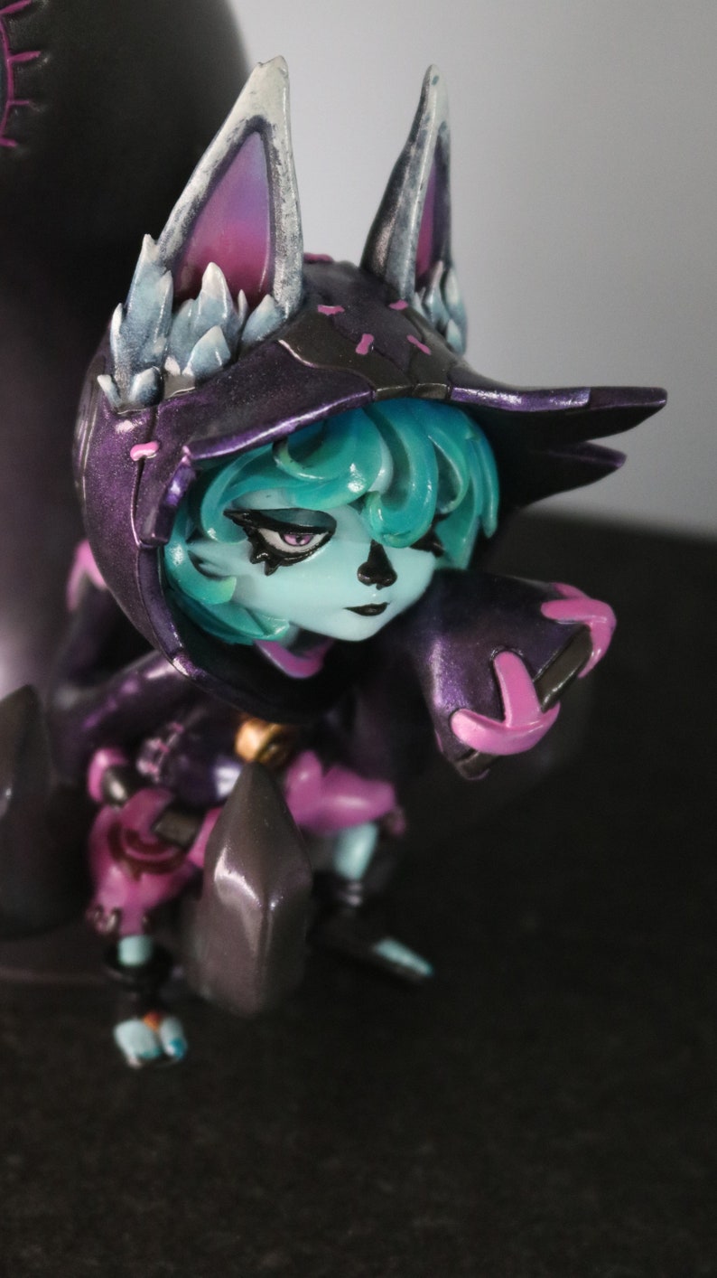 May include: A detailed figurine of a character with a fox-like hood and ears. The figure has teal hair, a pale blue face, and is dressed in a dark purple outfit with pink accents. The ears are white with purple and blue details.