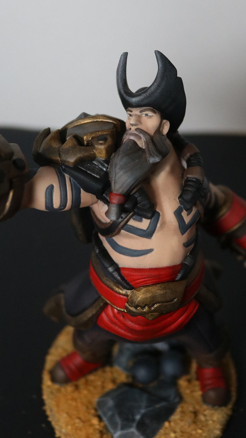 May include: A detailed figurine of a muscular character with a beard and a black hat. The figure is painted with a combination of colors, including red, gold, black, and tan. The character has tattoos and is posed in a dynamic stance.