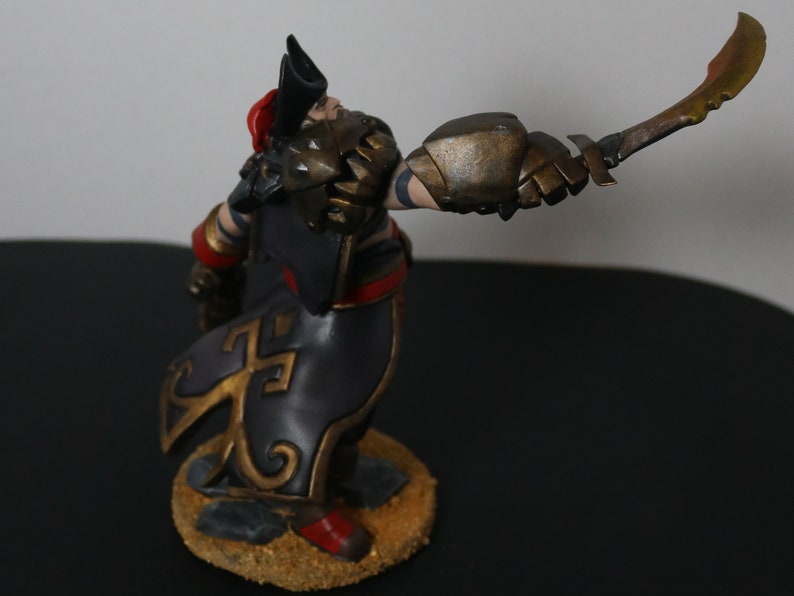 May include: A detailed figurine of a warrior in a dynamic pose, holding a curved blade. The figure is clad in armor with gold and bronze accents, a black cloak with gold designs, and a red hat. The base is textured with a sandy appearance.