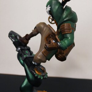 Pu&ograve; includere: Statuetta dettagliata di un personaggio in un abito verde con cappuccio e dettagli marroni, in posa dinamica. La figura &egrave; montata su una base verde scuro con una superficie strutturata simile alla sabbia, con estetica steampunk.