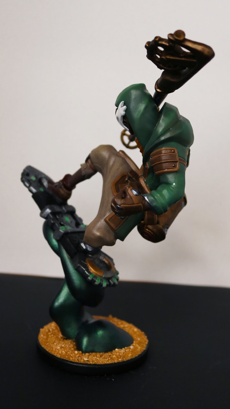 Pu&ograve; includere: Statuetta dettagliata di un personaggio in un abito verde con cappuccio e dettagli marroni, in posa dinamica. La figura &egrave; montata su una base verde scuro con una superficie strutturata simile alla sabbia, con estetica steampunk.