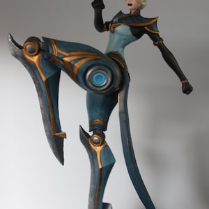 May include: Figurine of a female character in a dynamic pose, with one leg raised. The figure is predominantly teal and gold, with a white hairstyle and black accents. The base is a dark grey, textured design.