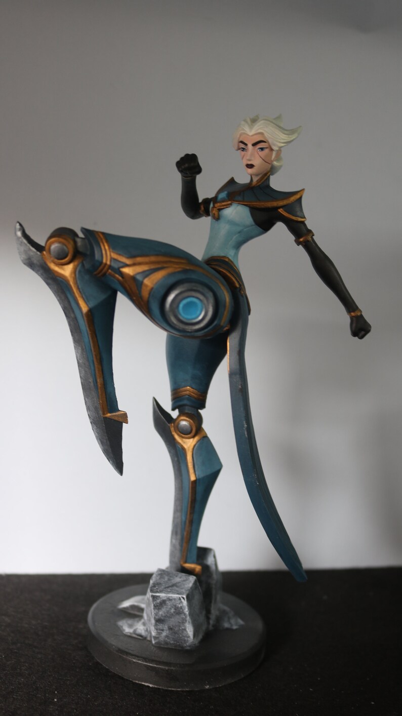 May include: Figurine of a female character in a dynamic pose, with one leg raised. The figure is predominantly teal and gold, with a white hairstyle and black accents. The base is a dark grey, textured design.
