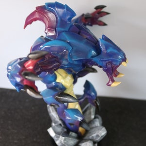 May include: A detailed figurine of a fantasy creature with a blue and purple armored body, yellow accents, and sharp claws. The figure is posed on a rocky base, showcasing intricate details and vibrant colors.