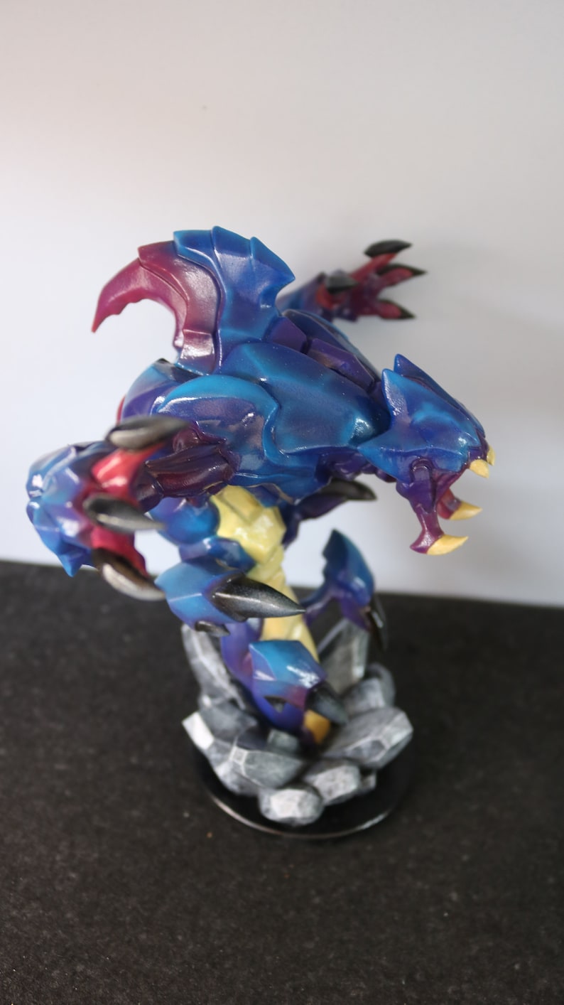 May include: A detailed figurine of a fantasy creature with a blue and purple armored body, yellow accents, and sharp claws. The figure is posed on a rocky base, showcasing intricate details and vibrant colors.