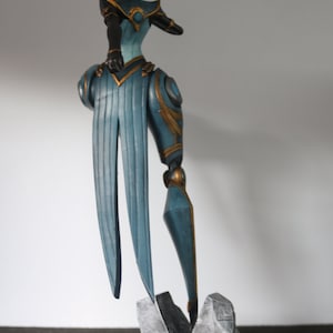 May include: A detailed figurine of a character in a teal and gold outfit, standing on a grey rock base atop a black circular platform. The figure has a stylized design with long, flowing elements and a distinctive hairstyle.