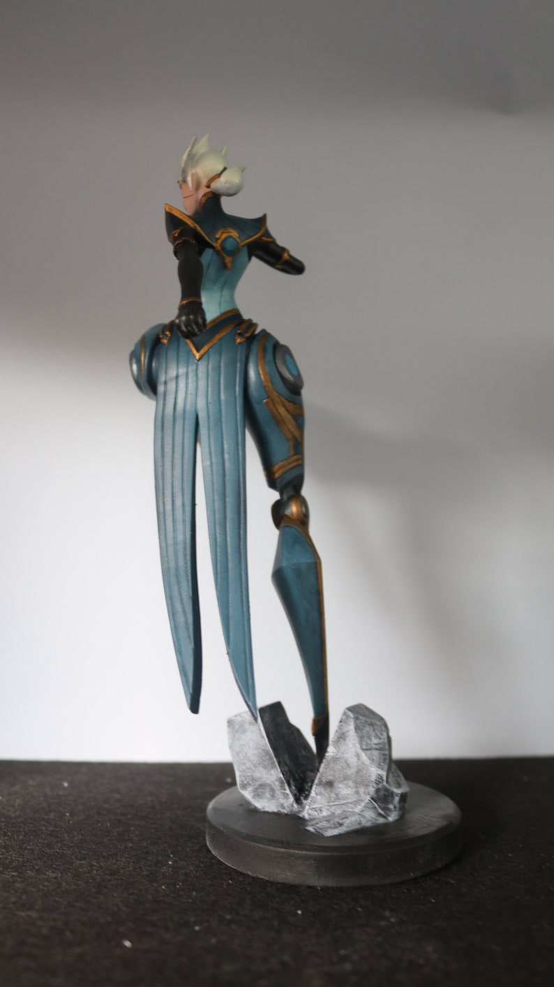 May include: A detailed figurine of a character in a teal and gold outfit, standing on a grey rock base atop a black circular platform. The figure has a stylized design with long, flowing elements and a distinctive hairstyle.