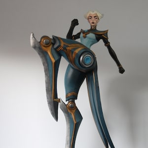 May include: A teal and gold figurine of a female character in a dynamic pose. The figure has short white hair and is wearing a teal and gold outfit. The figure is mounted on a black base.