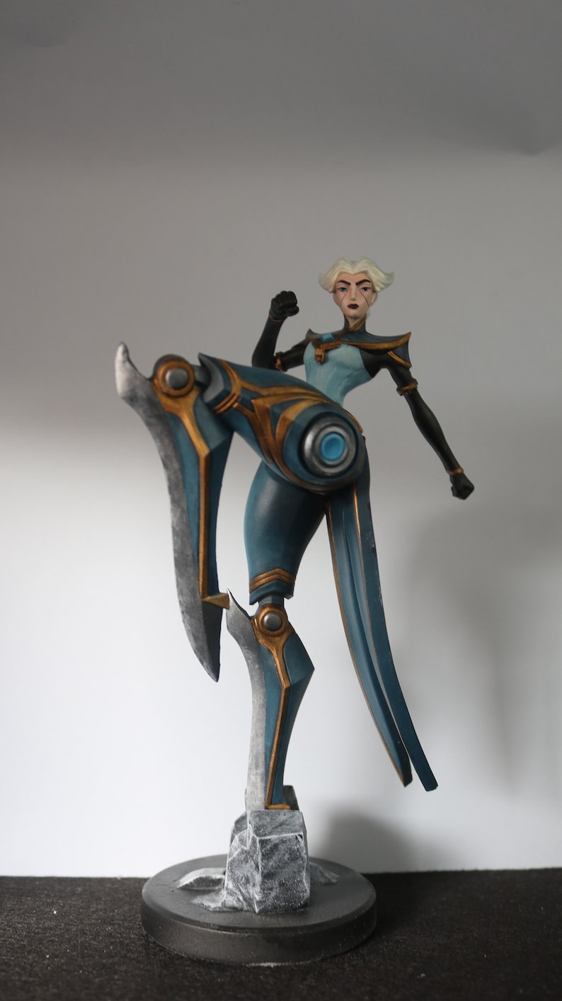 May include: A teal and gold figurine of a female character in a dynamic pose. The figure has short white hair and is wearing a teal and gold outfit. The figure is mounted on a black base.