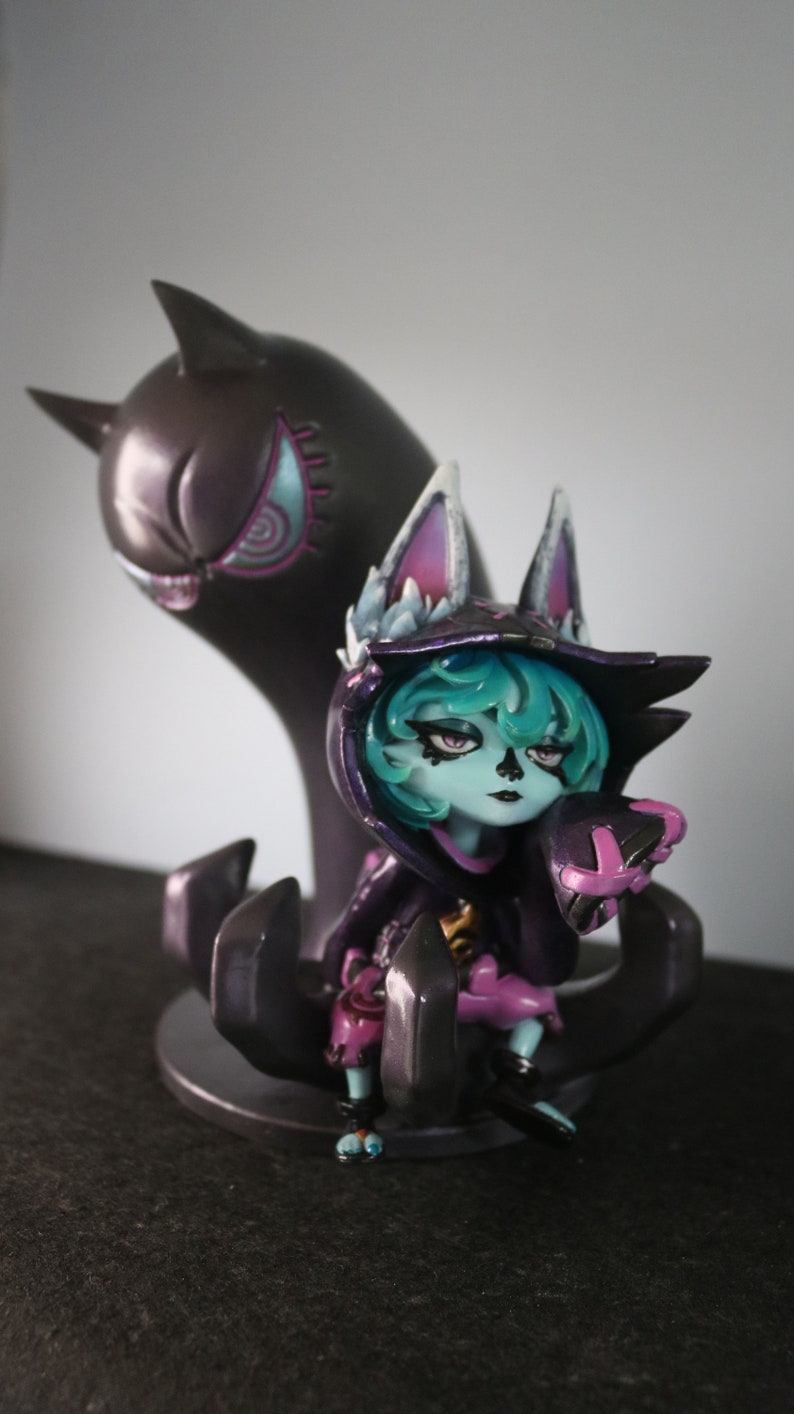 May include: A detailed figurine of a character with teal hair, wearing a black hooded outfit with purple accents. A dark purple ghost-like figure stands behind the character. The figurine is on a dark base.