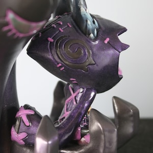 May include: A dark purple figurine with a stylized design. The figure has a horned head, stitched details, and a seated pose. Accents of pink and blue add contrast to the dark colour scheme. The figurine appears to be a fantasy or gothic-themed collectible.
