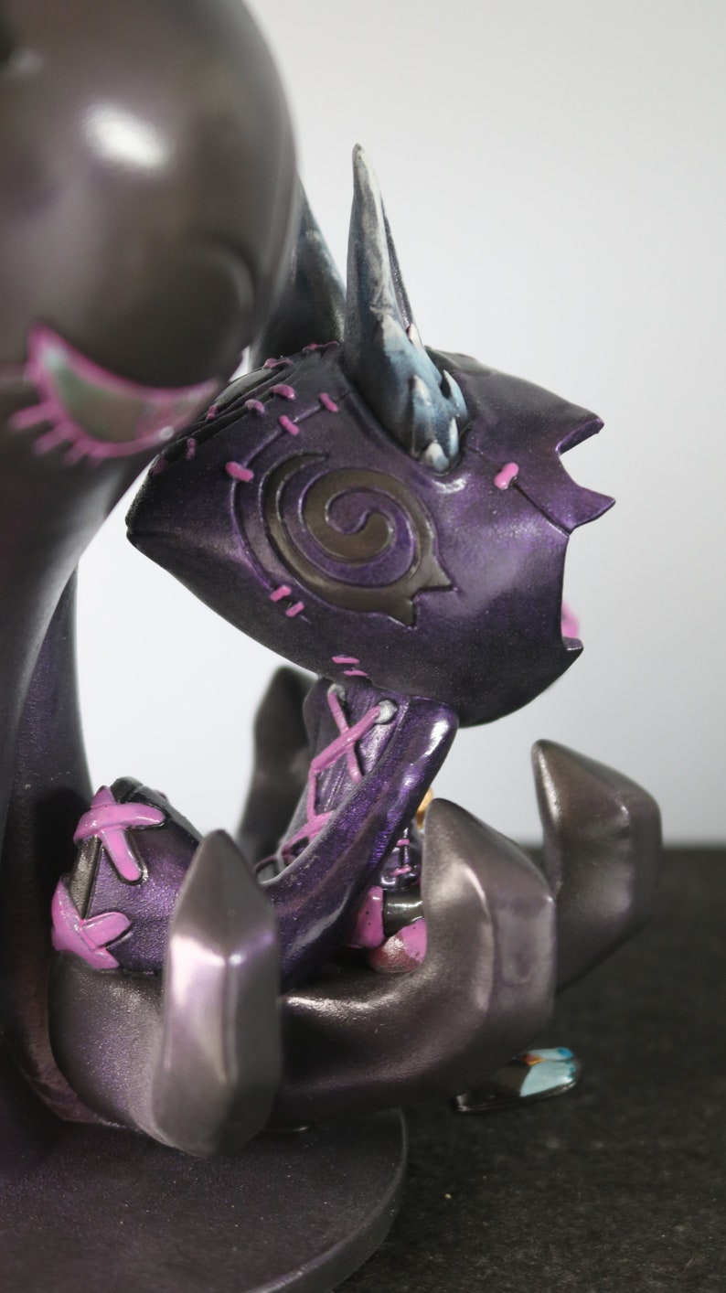 May include: A dark purple figurine with a stylized design. The figure has a horned head, stitched details, and a seated pose. Accents of pink and blue add contrast to the dark colour scheme. The figurine appears to be a fantasy or gothic-themed collectible.