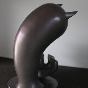 May include: A dark grey, abstract sculpture with a curved body and two small, pointed horns. The sculpture sits on a circular base and has a smooth, metallic finish. The object is likely a decorative piece or art object.