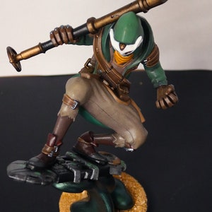 Pu&ograve; includere: Statuetta dettagliata di un personaggio in posa dinamica, che tiene un bastone. La figura indossa un cappuccio verde, pantaloni beige e stivali marroni. La base &egrave; una piattaforma circolare strutturata con una combinazione di colori verde scuro e oro.