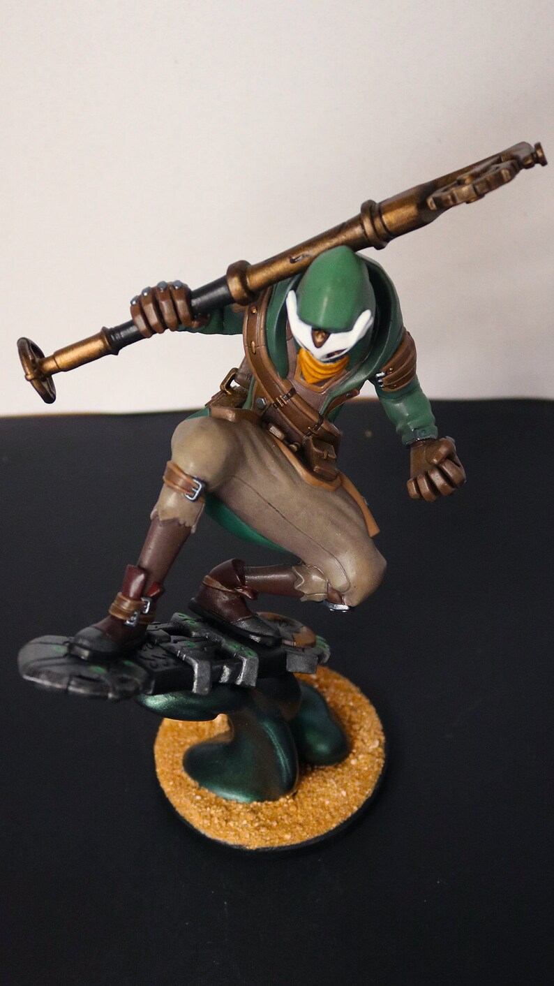 Pu&ograve; includere: Statuetta dettagliata di un personaggio in posa dinamica, che tiene un bastone. La figura indossa un cappuccio verde, pantaloni beige e stivali marroni. La base &egrave; una piattaforma circolare strutturata con una combinazione di colori verde scuro e oro.