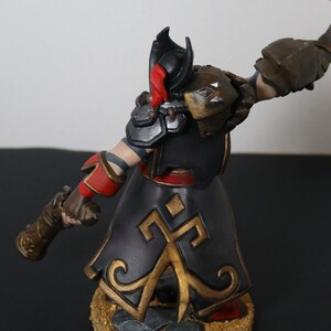 May include: A detailed figurine of a warrior in a black and gold outfit with red accents. The figure is holding a weapon and has a helmet with a red plume. The armor has silver and gold details. The base is textured.