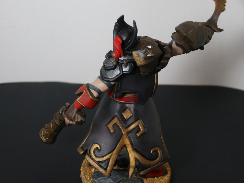 May include: A detailed figurine of a warrior in a black and gold outfit with red accents. The figure is holding a weapon and has a helmet with a red plume. The armor has silver and gold details. The base is textured.