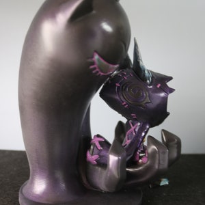 May include: A dark purple figurine of a creature embracing another. The larger figure has cat-like ears and a smooth finish. The smaller figure is held in the larger one's arms, with purple and pink accents.