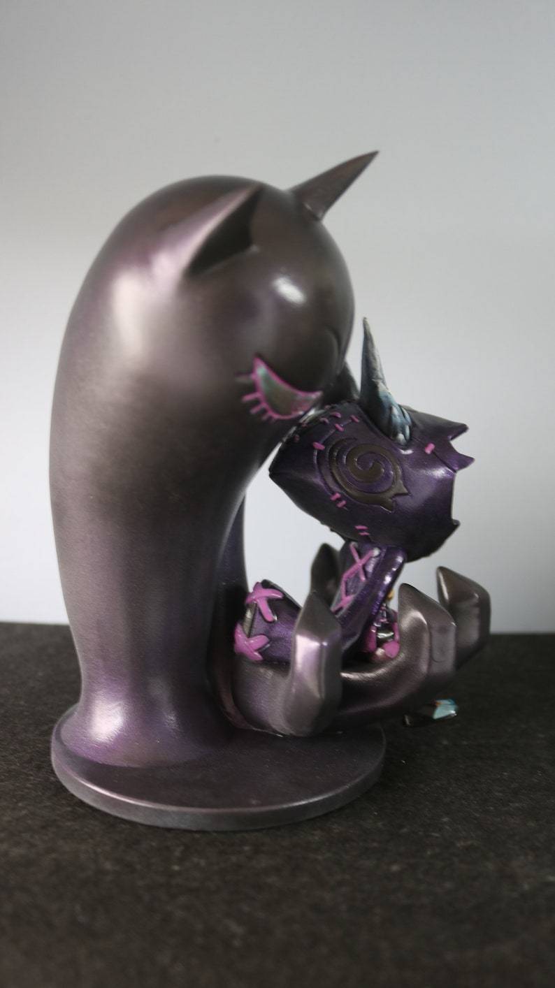May include: A dark purple figurine of a creature embracing another. The larger figure has cat-like ears and a smooth finish. The smaller figure is held in the larger one's arms, with purple and pink accents.