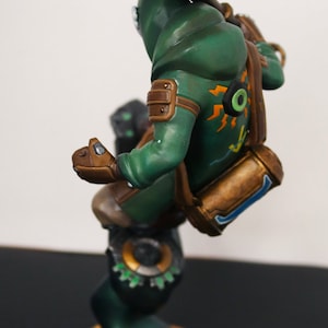 Pu&ograve; includere: Statuetta dettagliata di un personaggio in un completo verde con cappuccio, che tiene in mano un'arma. La figura ha dettagli marroni, uno zaino ed &egrave; montata su una base circolare con una superficie strutturata. La figura ha dettagli arancioni e gialli.