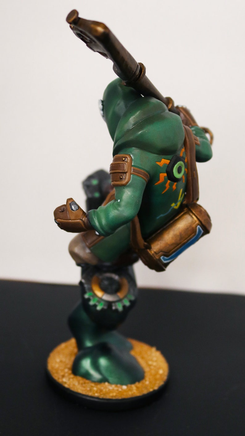 Pu&ograve; includere: Statuetta dettagliata di un personaggio in un completo verde con cappuccio, che tiene in mano un'arma. La figura ha dettagli marroni, uno zaino ed &egrave; montata su una base circolare con una superficie strutturata. La figura ha dettagli arancioni e gialli.