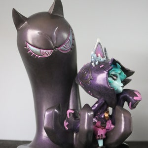 May include: A detailed figurine of a purple creature with cat-like ears and a hooded cloak, positioned next to a tall, dark purple figure with cat ears and stylized eyes. The figurine has teal skin and pink accents.