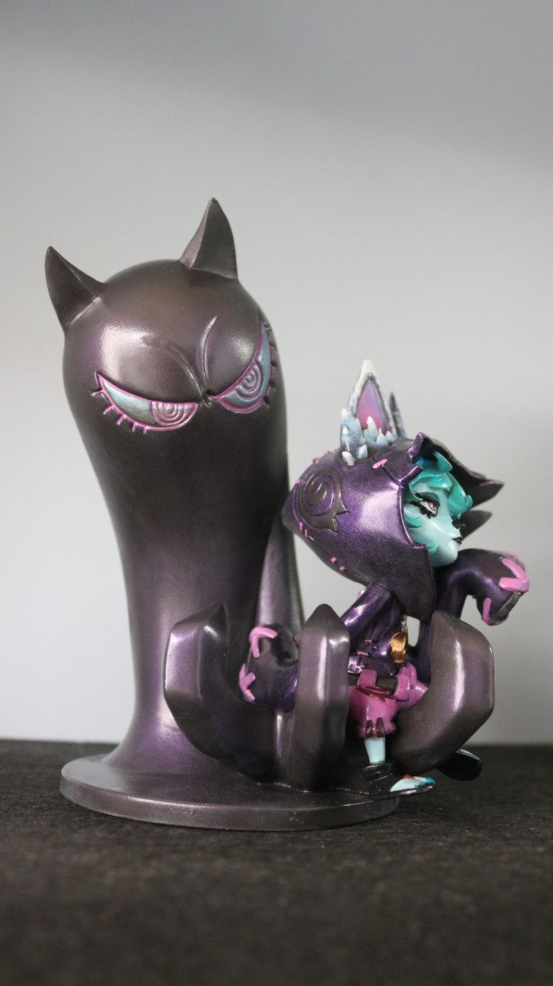 May include: A detailed figurine of a purple creature with cat-like ears and a hooded cloak, positioned next to a tall, dark purple figure with cat ears and stylized eyes. The figurine has teal skin and pink accents.